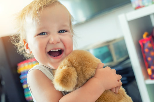 Cute Adorable Caucasian Blond Toddler Boy Having Fun, Laughing And Hugging Soft Puppy Toy Indoors. Cheerful Child Playing With Teddy Bear At Home. Kid Friend Animal Toy. Happy Childhood Concept