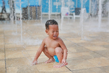 Happy little Asian baby boy having fun on water stream of a sprinkler. Kid playing in playground fountain in aqua park.