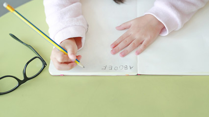 Close-up hands of little child writes ABC in a book or notebook with pencil on table in classroom against white background.