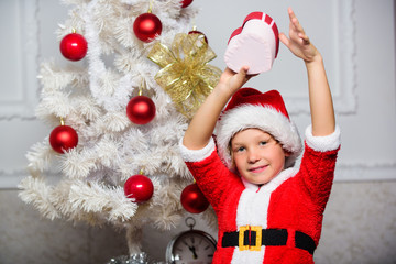 Christmas santa claus costume for child. Christmas party with santa costume. Christmas tree ideas for kids. Boy kid dressed as santa with red hat hold gift box near christmas tree