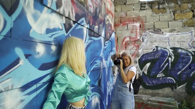 Blond girl poses for a girl photographer at the wall with graphitti in the summer in the city. Beautiful young girl model posing for a girl photographer. Graffiti wall. 4k