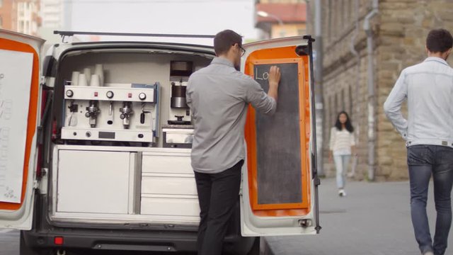Knee-up arc shot of young Caucasian man standing next to mobile coffee van in city street, writing menu on chalkboard on rear door, and casually dressed pedestrians walking by