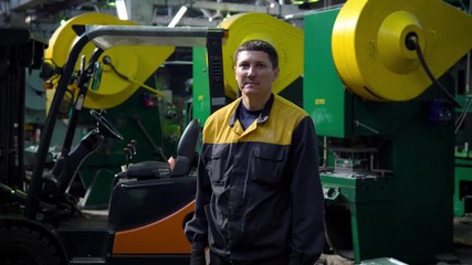 portrait of young employee in uniform of the enterprise, plant, warehouse on background of loader. He looks at camera and smiling