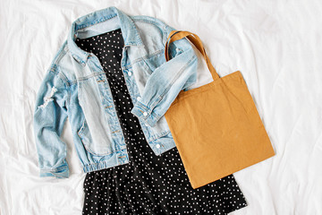 Blue jean jacket and  black dress with tote bag on white bed. Women's stylish autumn or spring  outfit. Trendy clothes. Flat lay, top view.