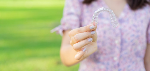 close up young beautiful asian woman smiling with hand holding dental aligner retainer (invisible) at outdoor nature park and garden background for beautiful teeth and dental treatment course concept
