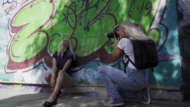 Blond girl poses for a girl photographer at the wall with graphitti in the summer in the city. Beautiful young girl model posing for a girl photographer. Graffiti wall. 4k