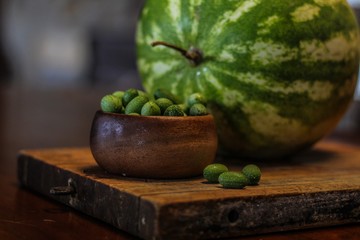 Still Life of a Large Watermelon on a Wooden Board next to a small wooden bowl of Mexican Sour Gherkins, selective focus on foreground