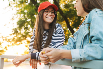 Image of two happy girls smiling and talking while bending on railing in green park