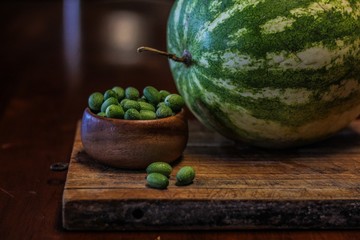 Still Life of a Large Watermelon on a Wooden Board next to a small wooden bowl of Mexican Sour Gherkins, selective focus on foreground