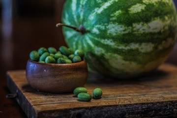 Still Life of a Large Watermelon on a Wooden Board next to a small wooden bowl of Mexican Sour Gherkins, selective focus on foreground