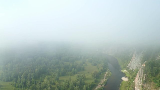Flight of the copter through the fog on a river valley in the early morning. Top view of the river and forest. To the right is a mountain range with steep cliffs.