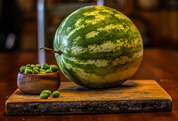 Still Life of a Large Watermelon on a Wooden Board next to a small wooden bowl of Mexican Sour Gherkins, selective focus on foreground