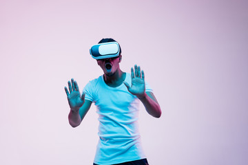 Young african-american man's playing and using VR-glasses in neon light on gradient background. Male portrait. Concept of human emotions, facial expression, modern gadgets and technologies.