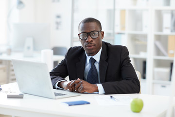Fototapeta na wymiar Portrait of African businessman in eyeglasses and black suit sitting at his workplace in front of laptop and looking at camera at office