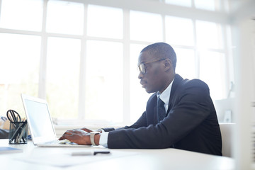 Concentrated African businessman in eyeglasses typing on laptop keyboard while sitting at his workplace at office