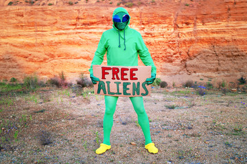 a man dressed in a green alien carnival costume suit at a lone rally with a sign "free aliens" on the background of a desert canyon