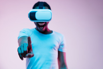 Young african-american man in VR-glasses in neon light on gradient background. Male portrait. Concept of human emotions, facial expression, modern gadgets and technologies. Pointing empty search bar.