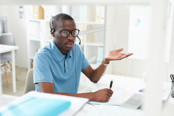 Naklejka na ściany i meble African young man in casual clothing sitting at the table wearing headphones and explaining something on the phone he working in a call center