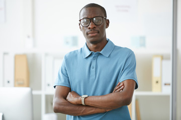 Portrait of African confident office worker in eyeglasses and in blue shirt standing with arms crossed and looking at camera at office