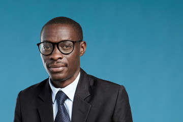Portrait of young African businessman in eyeglasses and suit looking at camera isolated on blue background