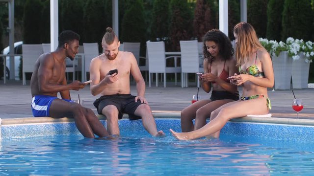 Happy multiracial young people communicating and laughing while viewing photos on cellphones sitting on edge of swimming pool with feet in water. Smiling diverse friends spending leisure in spa hotel