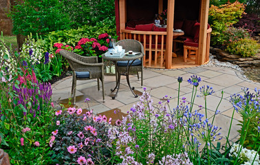The patio area with seating in an aquatic garden with colourful flower border with dahlia, agapanthus, salvia, phlox, foxgloves and hydrangeas in front of a Summer House
