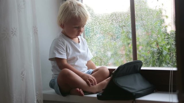 Adorable little boy, sitting on window shield, playing on tablet on a rainy day summertime