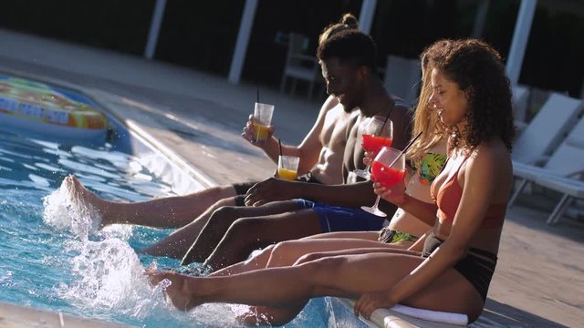 Group of happy multinational young people having fun sitting on edge of pool dangling legs and hitting water. Smiling diverse friends in swimsuits enjoying cocktails during summer leisure in hotel