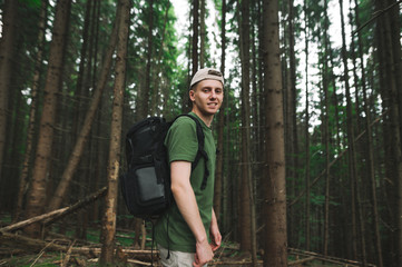 Portrait of an attractive young smiling tourist man looking at the camera walking in the mountain fir forest. Handsome relaxed careless male traveler posing on the camera at the woodland background.