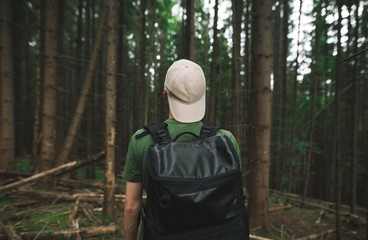 Fototapeta na wymiar Back view on a male hiker with a backpack in the cap standing in the mountain coniferous forest looking ahead. Tourist guy in green T-shirt standing among the fir trees on the woodland background.