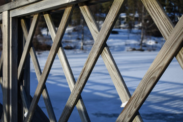 Obraz premium Criss cross pattern of a bridge's fence with forest and frozen lake in the background.