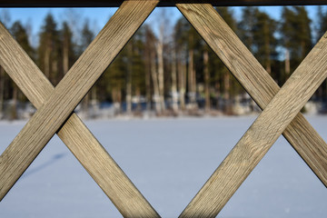 Criss cross pattern of a bridge's fence with forest and frozen lake in the background.