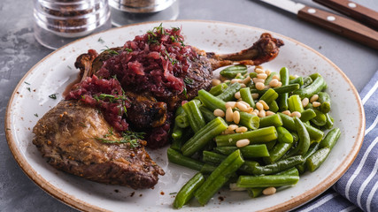 Juicy baked duck legs with green haricot and pine nuts on a plate served with fork and knife....