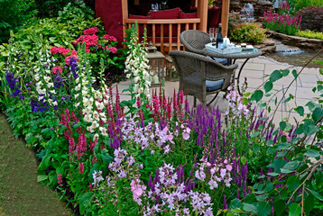 The patio area with seating in an aquatic garden with colourful flower border with salvia, phlox, foxgloves and hydrangeas in front of a Summer House