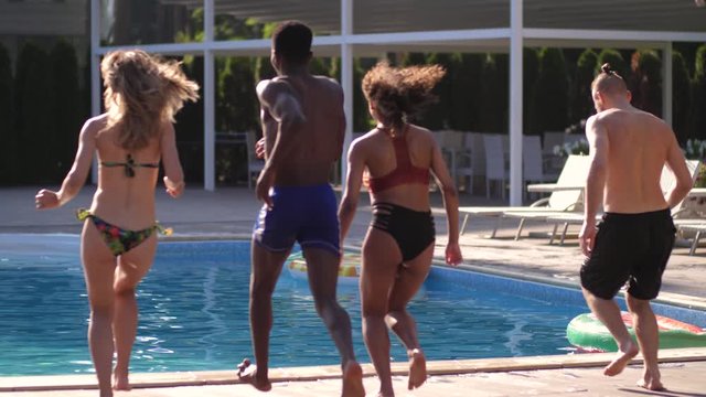 Back view of running multinational friends jumping into water of swimming pool and making a splash. Playful multiracial young people spending summer leisure by outdoor pool of luxury spa hotel
