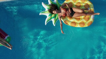 Drone view of diverse happy young women relaxing on colorful inflatable pineapple and watermelon in swimming pool of luxury spa hotel. Aerial shot of female friends enjoying leisure on pool floats