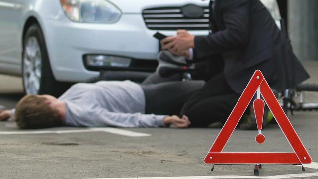 Male driver calling ambulance, unconscious bicyclist asphalt, road danger sign