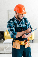 happy bearded worker in safety helmet writing while holding clipboard