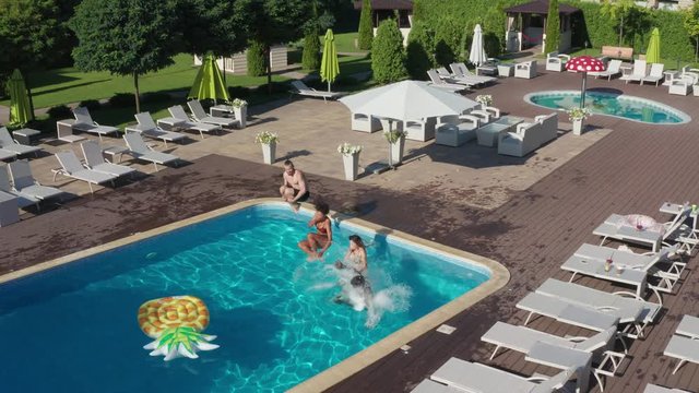 Drone view of playful multinational friends holding hands running and jumping into clear blue water of swimming pool. Happy diverse young people having fun spending summer holidays in luxury spa hotel
