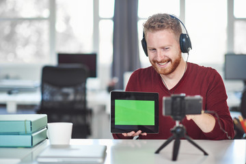 Handsome Caucasian employee having presentation over the internet. In hands is tablet and on head are headphones. Start up business concept.