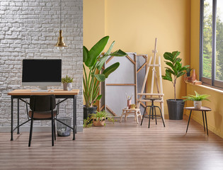 Modern yellow wall background and white brick wall style, wooden working table computer style, easel painting decoration vase of plant.