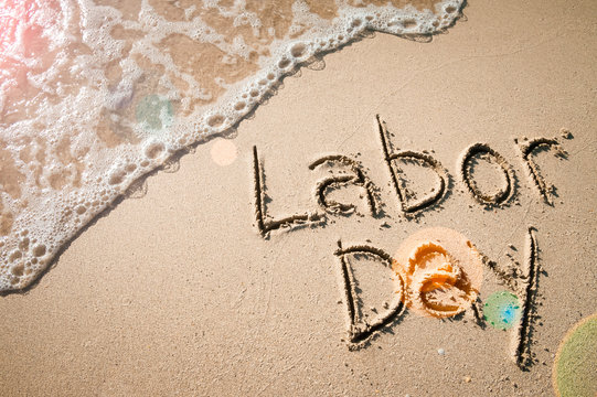 Labor Day message handwritten on the smooth sand of an empty beach with sunny lens flare across an oncoming wave