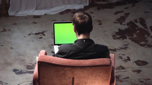 View from the back of young man in black shirt sitting in the brown comfortable chair and working on laptop computer with chroma key green screen. Stock footage. Place of your own content.