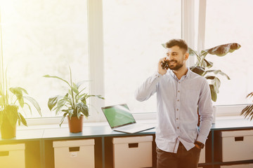 Portrait of a cheerful smiling men office worker talking on mobile phone while standing in modern...