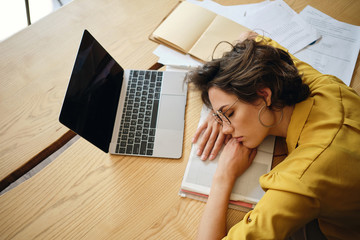 Top view of young tired woman dreamy asleep on desk with laptop and documents under head at...