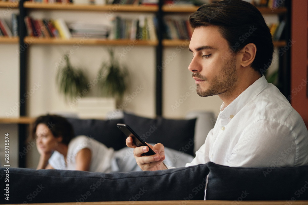 Poster side view of young handsome businessman sitting on sofa with colleague on background while thoughtfu
