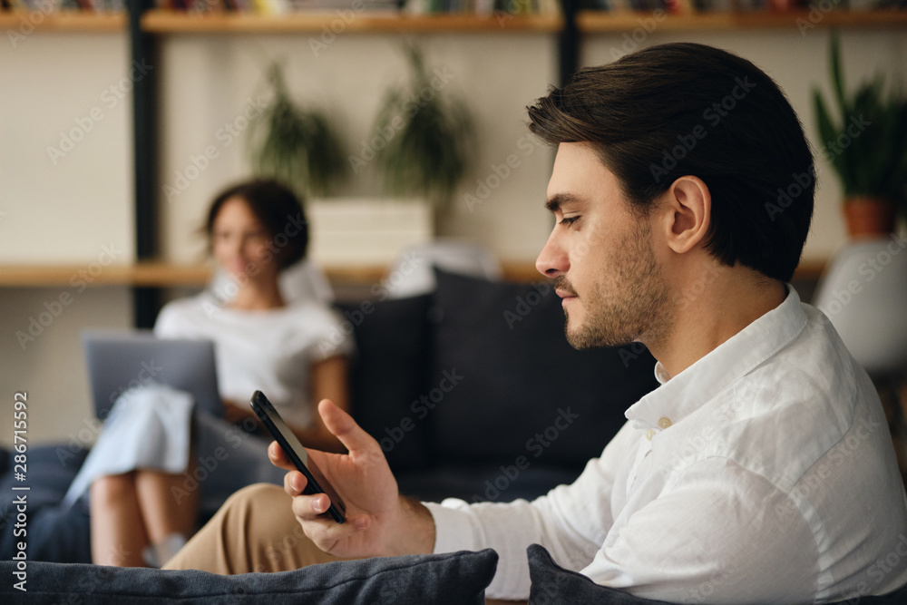 Poster Side view of young handsome businessman sitting on sofa with colleague on background while dreamily using cellphone in modern co-working space