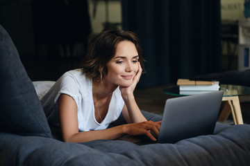 Young attractive smiling woman lying on sofa happily working on new project with laptop in office