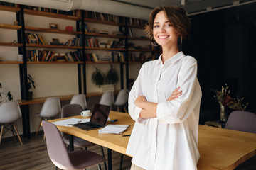 Young cheerful woman in white shirt happily looking in camera with desk on background in office