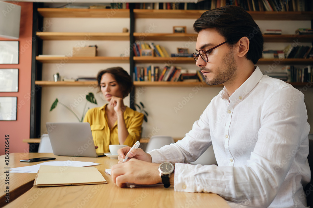 Poster Young attractive man in eyeglasses sitting at the table thoughtfully writing in notepad with colleague on background at work in office